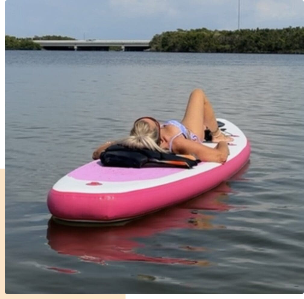 Person relaxing on a pink paddleboard in calm water near a bridge.