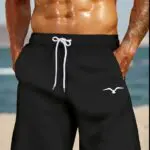 Man wearing black swim shorts with a white drawstring, standing at the beach.