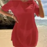 Person in a red dress standing on a sandy beach with rocks and ocean in the background.