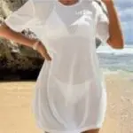Person in a white dress standing on a sandy beach near rocks and ocean.